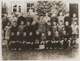 Three rows of boys line up for a school photo. A small chalkboard sits on top of one of the boy’s feet in the center of the photo. No text is visible.