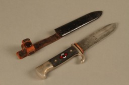 This Hitler Youth knife and case is shaped like a military bayonet and bears the emblem of the Hitler Youth.
Beginning in 1933, the Hitler Youth and the League of German Girls had an important role to play in the new Nazi regime. Through these organizations, the Nazi regime planned to indoctrinate young people with Nazi ideology. This was part of the process of Nazifying German society. The aim of this process was to dismantle existing social structures and traditions. The Nazi youth groups were about imposing conformity. Youth throughout Germany wore the same uniforms, sang the same Nazi songs, and participated in similar activities. Knives such as this one emphasize the paramilitary nature of the Hitler Youth organization. It was designed to train boys as future fighters and soldiers for the Nazi cause.
These items were found in a warehouse in Aachen, Germany, by US Army soldier Sidney Conners in July 1944. 