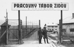 Two uniformed soldiers stand in front of a half-open gate. The wooden gate behind them has barbed wire along the top.There is a large sign above the gate. A couple of buildings can be seen behind the gate.