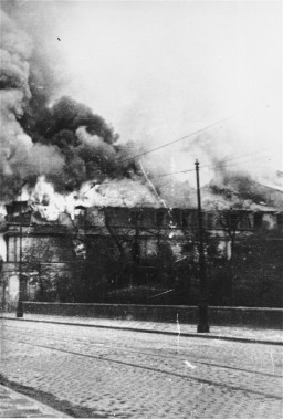 Photograph of a large building engulfed in flames taken from across the street.