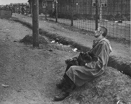 An emaciated victim sits on the ground outside of a fenced area. They are wearing a long coat and leaning back with a look of anguish on their face. The victim appears to be holding something in their lap. Several buildings can be seen behind the fence.