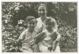 In a black and white photograph, a woman, Vilma Grunwald, sits in a garden with her baby son on her lap. Her older son, who is a young child, sits to her right. The family is posing for the camera.