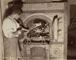 A man stands in front of an open brick crematorium oven holding a long handled fire iron. He is wearing a light long sleeved button up with the sleeves rolled and long pants. He has very short medium dark hair and light skin tone.