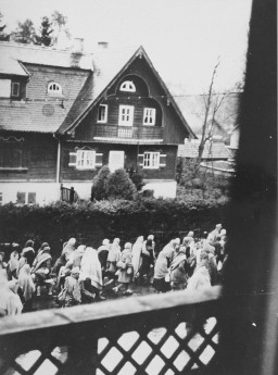 Photograph taken out of a small window, featuring groups of prisoners marching through a village.