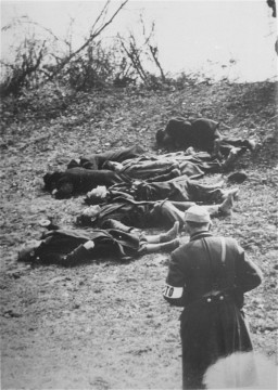 Several clothed corpses lay on the group of an elevated river bank. A uniformed soldier stands at the bottom of the bank with a rifle slung over his shoulder.