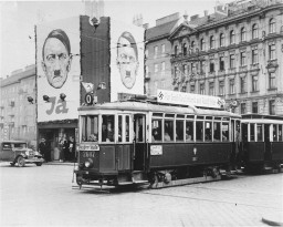 A streetcar decorated with swastikas passes billboards displaying Hitler's face. [LCID: 70073]
