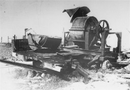 Large barrel like structure with a circular crank attached. It sits on what appears to be an abandoned trailer in the middle of a field.