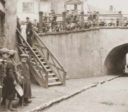 Several people stand along an elevated walkway leading towards a bridge. A wooden staircase has been added to the walkway, providing access to the road below. A few individuals stand along the side of the lower road.