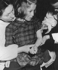 اقوام متحدہ کے اہلکار حراستی کیمپ میں زندہ بچ جانے والے 11 سالہ بچے کو حفاظتی ٹیکے لگا رہے ہیں جو آشوٹز کیمپ میں طبی تجربات کا شکار ہوا۔ برجن۔بیلسن کے بے دخل افراد کا کیمپ، جرمنی، مئی 1946 ۔