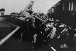 A large crowd of people squeeze in between two railroad cars. A soldier with a rifle over his shoulder can be seen perched atop one of the open-top railway cars.