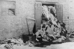 Garments of all shapes, sizes, and colors spill out of a doorway. The pile goes up to the ceiling of the brick warehouse and can be seen through the windows.