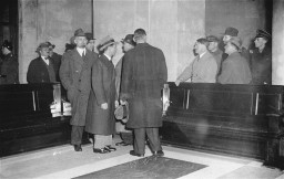 Adolf Hitler stands with his hand on the back of a long bench with Hermann Göring and Joseph Goebbels to his left at the site of the parliament building fire. 
Seven men stand in a group in front of the long bench, wearing coats and other outerwear.