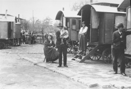 A family stands outside of their wagon while interned in a Zigeunerlager ("Gypsy camp"). In the background, children are crowded around Eva Justin. Justin worked for the Center for Research on Racial Hygiene and Demographic Biology. Schleswig-Holstein, Germany, 1938. 
During the Nazi era, Dr. Robert Ritter was a leading authority on the racial classification of people pejoratively labeled “Zigeuner” (“Gypsies”). Ritter’s research was in a field called eugenics, or what the Nazis called “racial hygiene.” Ritter worked with a small team of racial hygienists. Among them were Eva Justin and Sophie Ehrhardt. Most of the people whom Ritter studied and classified as Zigeuner were German Roma called Sinti. Sinti are a subgroup of Romani peoples. 
In 1935, municipal authorities across Nazi Germany began to force Romani families to move into Zigeunerlager (“Gypsy camps”). Eventually, these camps were centralized under the authority of the Nazi Kripo (criminal police). Ritter and his team regularly examined the individuals in these camps. They sometimes used threats or bribes to force people to cooperate. The creation of these camps was one of the Nazis’ early steps toward the genocide of Romani peoples. Many Romani victims were later deported to concentration camps, ghettos, or killing centers. 
Source Record ID: Bild 146/87/108/66