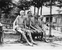 Four survivors sit on a bench outside of a wooden building. All of them have shaved heads and are wearing tied clothing as shorts. Most are barefoot, with their boots sat under the bench.