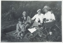From left to right, one child and three adults sit on a blanket in a wooded area with food and drink, and games around them. All share a medium-light skin tone.