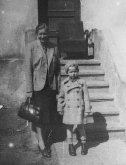 A woman with light hair and light skin tone smiles while holding a large bag in her right hand. A small boy with light skin tone stands next to her wearing a hat.
