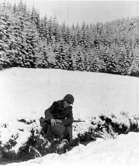 Un GI américain puise de l'eau dans un ruisseau avec son casque en acier. 22 décembre 1944. Corps de transmission de l'Armée américaine, photographie prise par J. Malan Heslop.