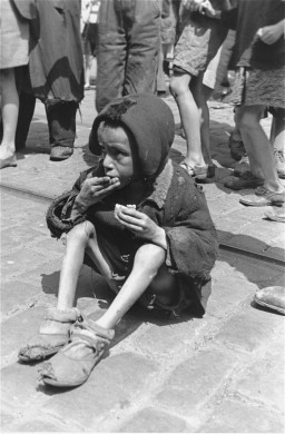 A severely emaciated child with medium-light skin tone sitting on a street eating while people walk around them. The child is wearing a heavy jacket and worn slippers with bare legs.