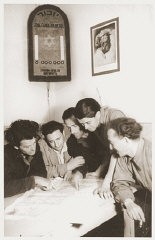 Miembros del kibbutz Nili (una cooperativa agrícola sionista) estudian un mapa de Palestina. En una pared se encuentra una placa que conmemora a los seis millones de judíos asesinados durante el Holocausto. En la otra pared, una fotografía del líder sionista socialista, Berl Katznelson.  Pleikershof, Alemania, 1945-1948.