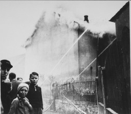Firefighters use water hoses to save a home while the building next door is covered in smoke and burns. A group of people of various ages stand off to the left and look directly at the camera.