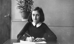 A young girl with shoulder length dark hair and light skin tone sits at a desk with writing materials.