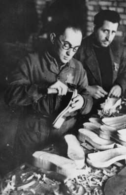 Two individuals stand at a work table and appear to be carving the bottom of shoes. The man closest to the camera is balding with dark hair and light skin tone. The man behind him has short dark hair and light skin tone. Shavings litter the table amongst stacks of materials and finished products.