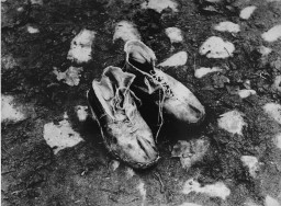A pair of muddy worn leather lace up shoes sit on the rocky ground.
