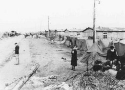 View of Bergen-Belsen concentration camp after the liberation of the camp. A row of small tents has been pitched outside the barracks.  A group of survivors huddles in front of one of the tents. 