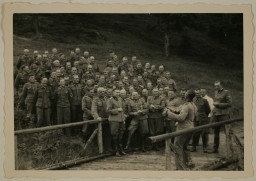 Dozens of unformed soldiers stand in a field looking towards a lone performer with a guitar. Several people appear to be swaying or singing along with the music. A foot bridge can be seen along the bottom of the photograph.