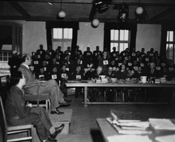 A witness testifies during the Mauthausen concentration camp trial. The man standing in the background is defendant Willy Eckert, a member of the SS. The trial took place before an American Military Tribunal in Dachau, Germany. March-May 1936. 