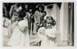 Two little girls with curly hair pulled up in updos and light skin tone stand among a group of people. They wear light colored dresses and are holding onto small bouquets and what appears to be a veil. A group of people gather in the background to watch the procession.