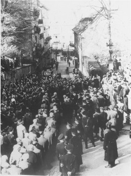 A large crowd watches 2 long lines of men walking down the center of the road. Uniformed men stand on either side of the lines supervising.