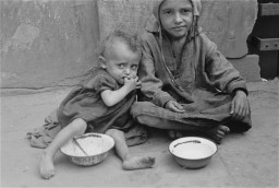 Two malnourished children sit in the dirt eating from shallow plates. The one on the left is younger, almost toddler age, with short curly hair and medium skin tone wearing a large dark dress. The child on the right is older, with medium skin tone,  a head covering, and baggy clothes.