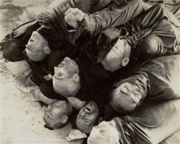 A pile of corpses, wrapped in fabric from the neck down. The faces and feet of the victims can be seen.
