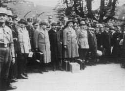 A group of men stand together. Most are wearing hats and outerwear. Uniformed guards stand around the perimeter of the group.