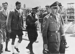 Heinrich Himmler in full uniform leading a group of other men down a path. Some men are in uniform, others are in suits. All men in the photo appear to have light skin tone and dark short hair.