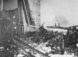 To the left, a fireman aims a water hose at a pile of smoldering rubble. There is a line of firemen standing behind him against a wall.
