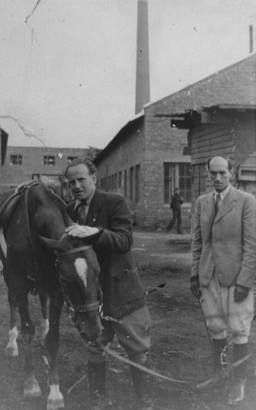 Two men look toward the camera wearing suits, riding boots and gloves. A horse in tack stands next to the man in the center. He leans in towards the horse with a hand on the horse’s head.