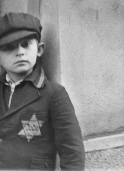 The cropped image of a young boy standing against the corner of a stone wall. The young boy wears a dark cap and jacket. On the left pocket of the jacket is a large six-pointed star, the Star of David, with the word “Jude” in the center. Only the top right half of the boy can be seen in this photograph.