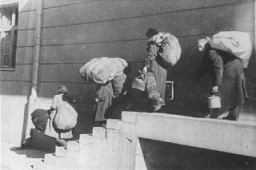 A family of Macedonian Jews carries their luggage down a flight of stairs as they leave the Tobacco Monopoly transit camp for the deportation trains. Skopje, Yugoslavia, March 1943.
