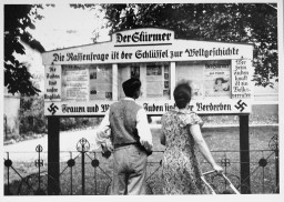 A large outdoor newspaper display covered in German writing. A couple with light skin tone stands in front of the display reading the displayed pages.