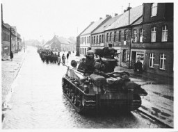 دبابة أمريكية خلف مواطنين ألمان من مدينة غاردليغن يسيرون إلى مخزن خارج المدينة حيث سيستخرجون أكثر من ألف جثة لأولئك السجناء الذين قُتلوا من قبل قوات الأمن الخاصة. 18 أبريل 1945.