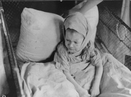 A small child with light skin tone lays in a hospital bed against a large pillow. They appear to wear a hospital gown and have a dressing wrapped around their head and under their chin. The sides of the hospital bed have fencing to prevent the child from rolling out.