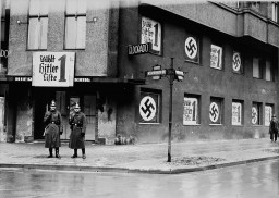 Two uniformed policemen stand outside of a shuttered nightclub. The windows and signage have been covered with boards featuring the swastika or German text.