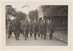 A group of uniformed Nazis walk down what appears to be a rural dirt road. There is a brick structure behind them, next to the road.