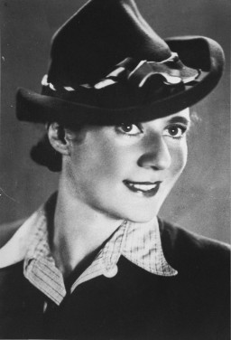 A woman with light skin tone smiles at the camera. She is wearing a collared shirt and a large fedora-type hat cocked to the side.