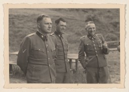 Three uniformed male officers with light skin tone stand in a field with picnic tables behind them. The farthest two officers are talking and laughing.