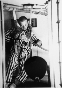 An individual with light skin tone  in a striped uniform hangs by a chest harness with various medical instruments attached in a mostly clear chamber. Their head has fallen forward.