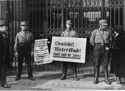 Three uniformed Storm Troopers with light skin tone stand in front of a closed shop. The soldier in the center holds a boycott sign in German, while another sign hangs from the gates covering the shop entrance. The soldier to the right appears to be speaking to a feminine figure with light skin tone in dark clothes.