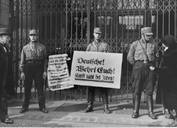 Fırtına Birlikleri’nin (SA) üyeleri boykot pankartlarıyla bir Yahudiye ait dükkâna müşterilerin girişini engelliyor. Pankartlardan birinde şu öneriliyor: "Almanlar! Kendinizi savunun! Yahudilerden alışveriş yapmayın!" Berlin, Almanya, 1 Nisan 1933.